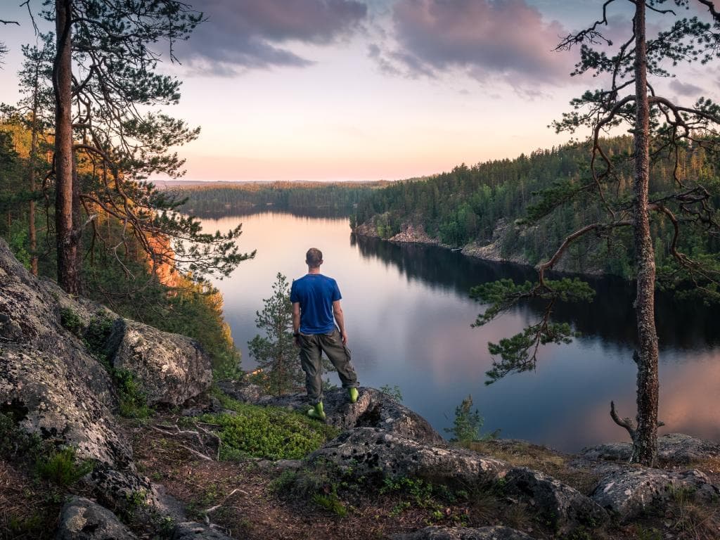 En man sitter på en höjd och blickar ut över en sjö och skog under solnedgång.