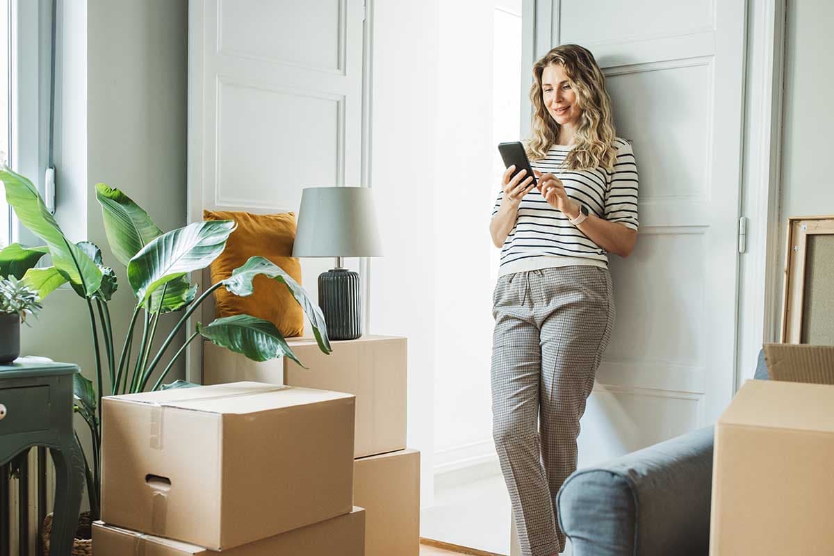 En kvinna lutar sig mot en dörrpost med mobilen i handen, omgiven av flyttkartonger i ett ljust rum.