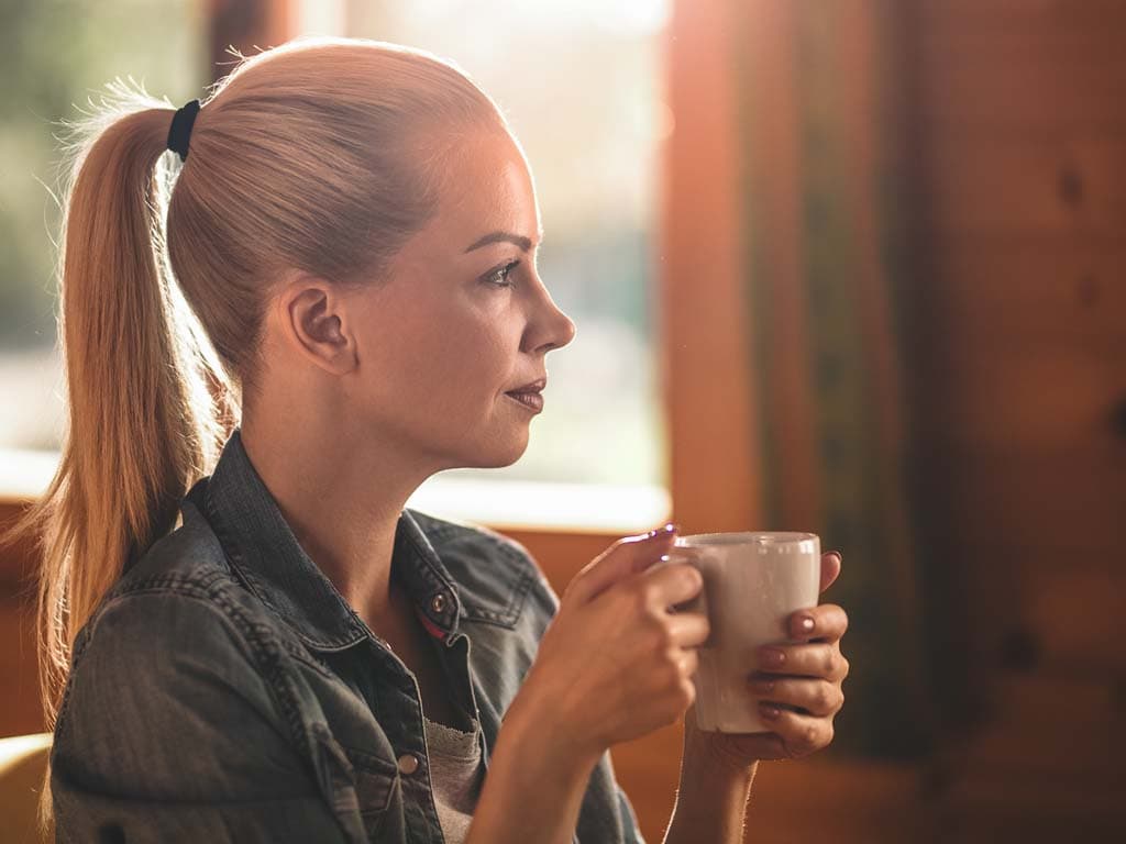 En kvinna håller en kopp och stirrar ut i fjärran, försjunken i tankar.