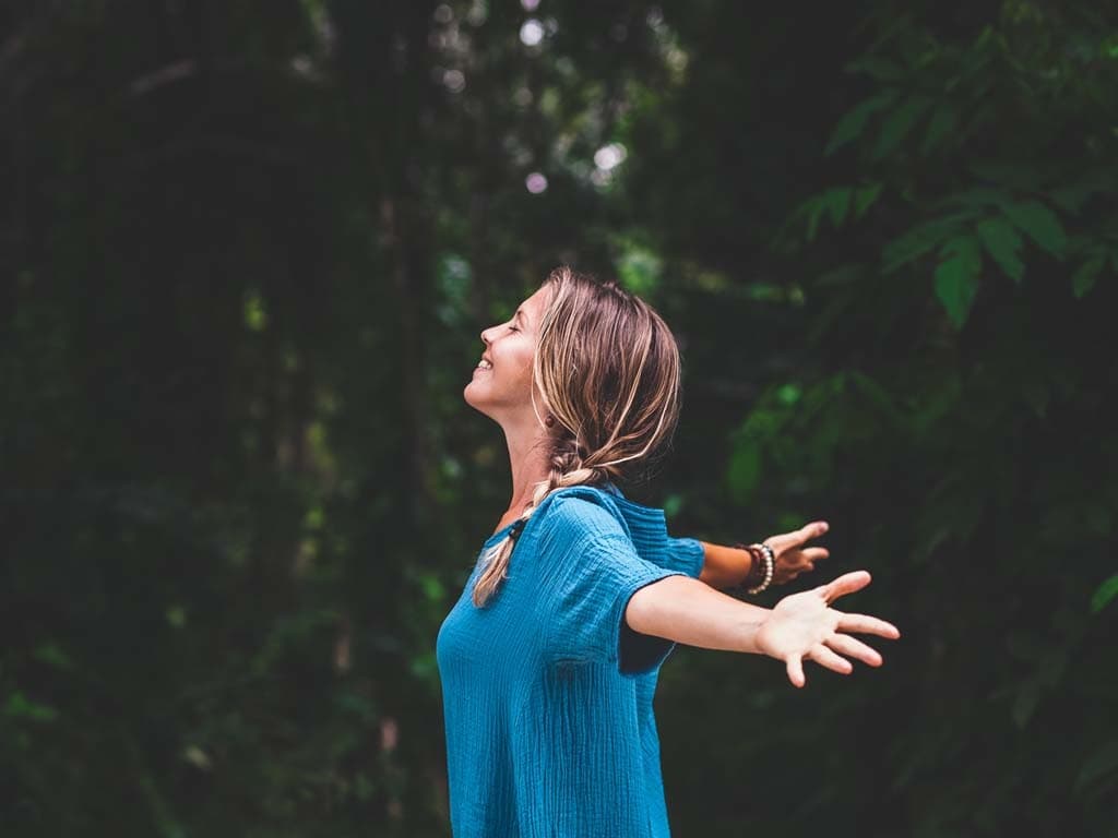 En kvinna står i naturen med armarna utsträckta och ögonen slutna, njuter av frisk luft.