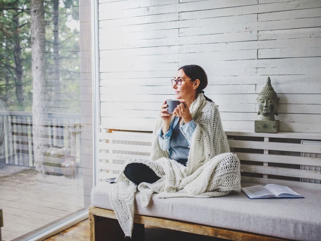 En kvinna sitter vid ett fönster i ett ljust rum och håller en kopp te, tittar ut mot naturen i ett lugnt ögonblick.