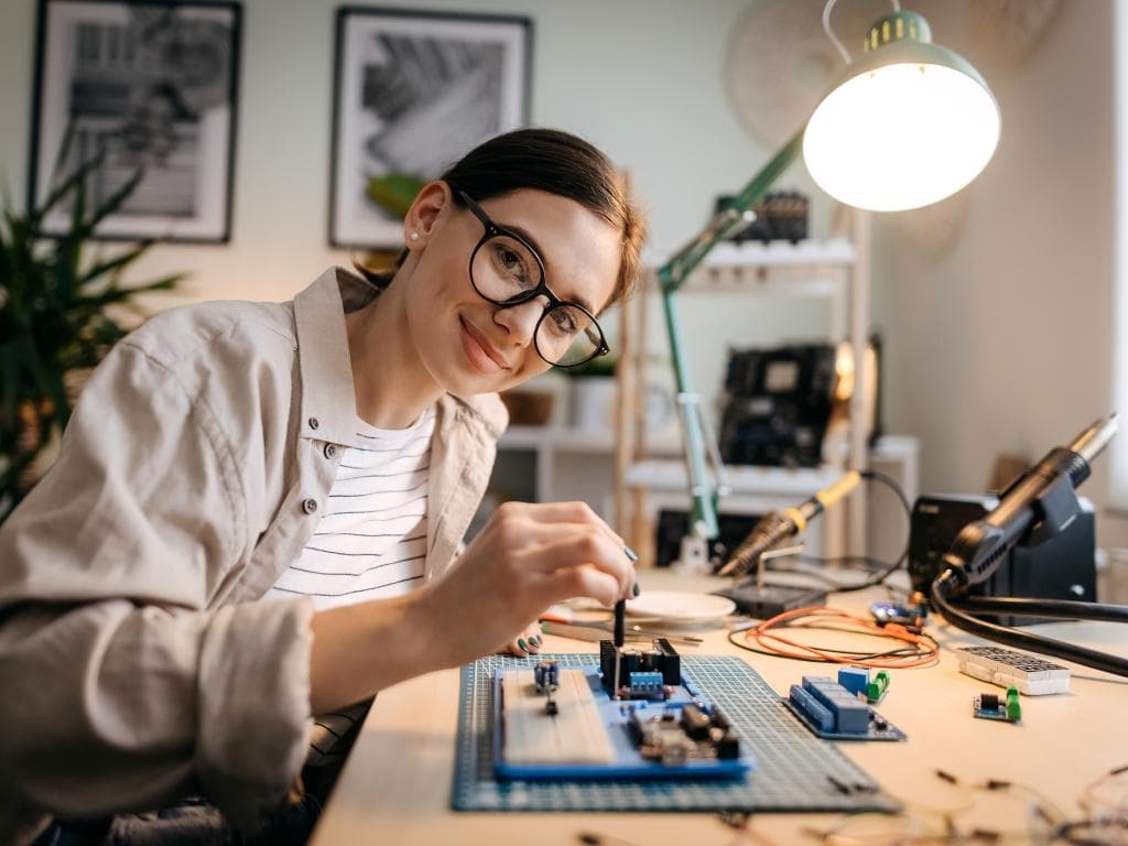 En kvinna arbetar fokuserat med elektronik vid ett skrivbord i hemmaverkstadsmiljö.