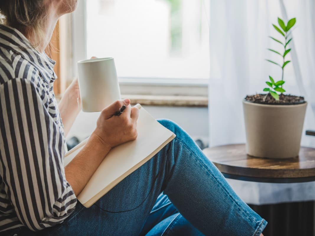 En kvinna sitter vid ett fönster med en kopp i ena handen och anteckningsblock i den andra.