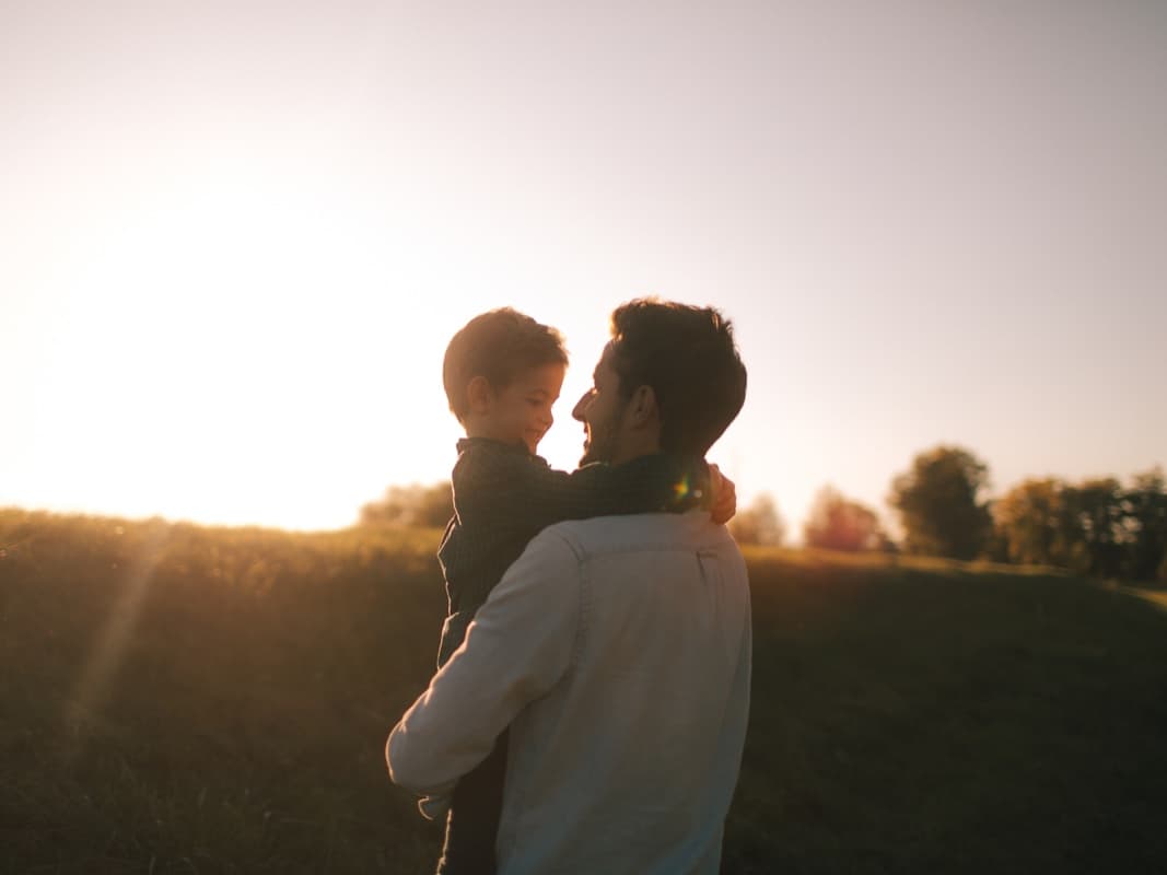En man håller ett barn i famnen ute i naturen vid solnedgången.