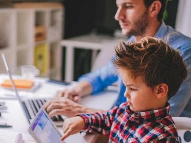 En kvinna arbetar vid dator hemma medan ett barn sitter bredvid, båda är fokuserade.