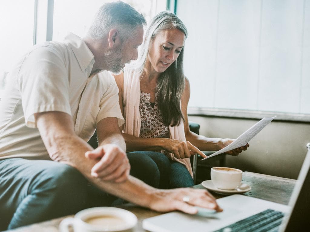 Ett par sitter i vardagsrummet och granskar papper och en laptop, diskuterar ekonomi.