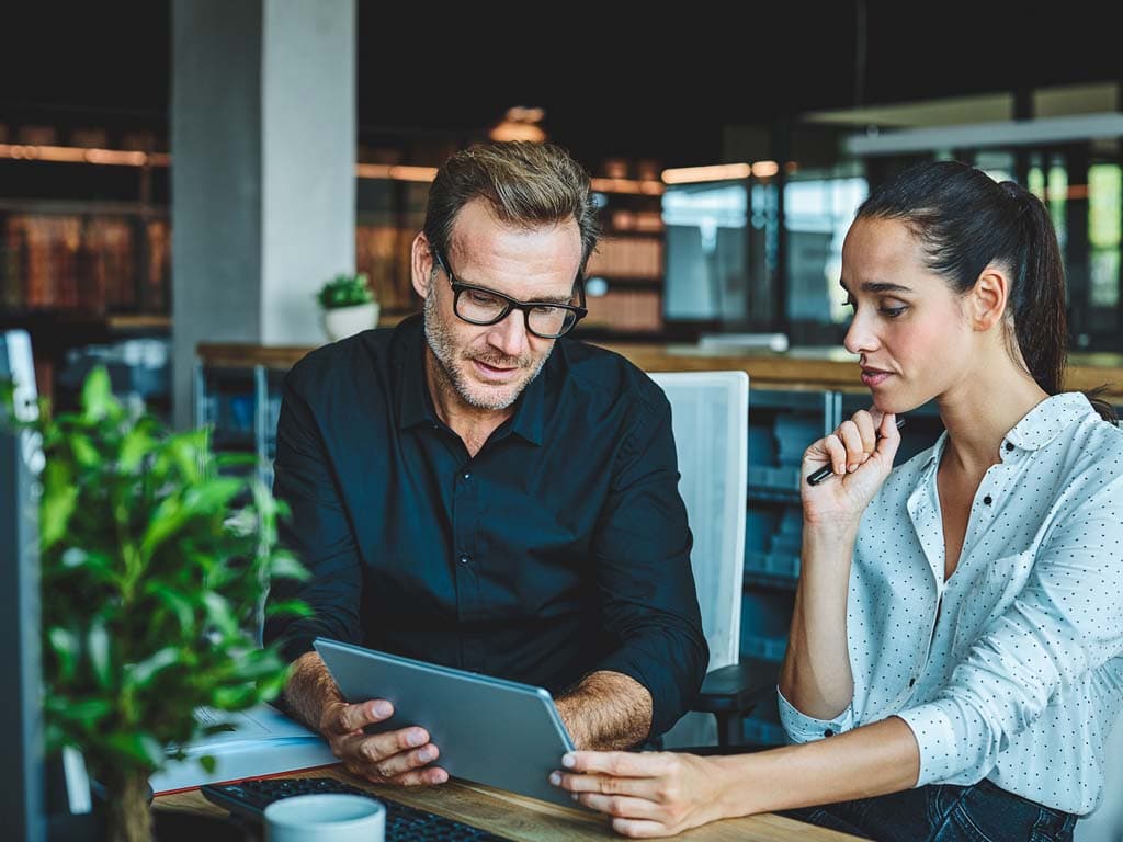Två kollegor sitter vid ett bord och tittar tillsammans på en surfplatta, leende.