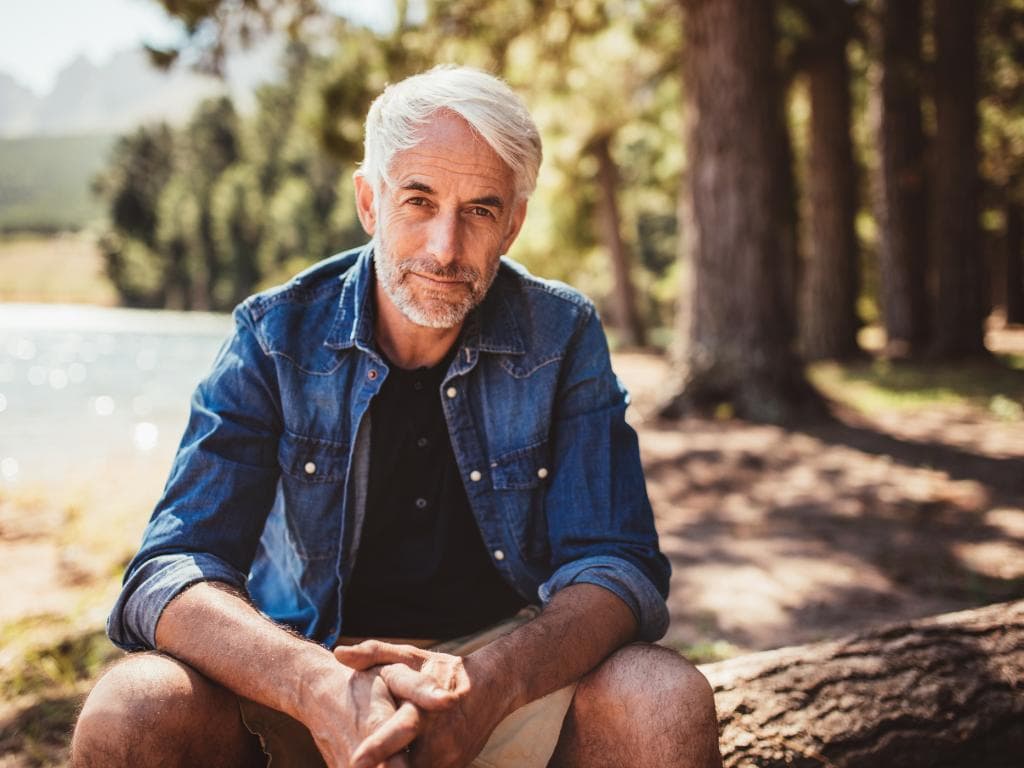 En äldre man sitter avslappnat på en bänk vid en sjö, omgiven av skog och solsken.