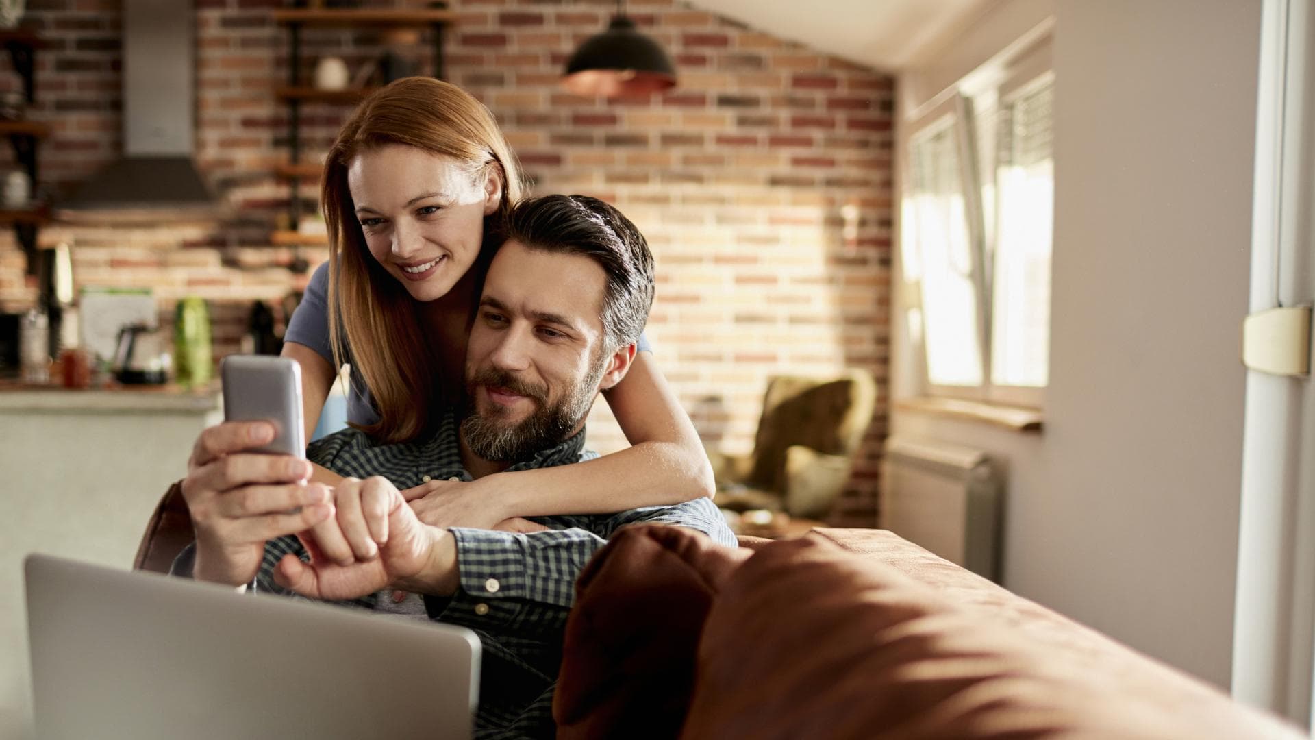 Ett par sitter tillsammans i ett mysigt vardagsrum och ler medan de tittar på något på mobilen.